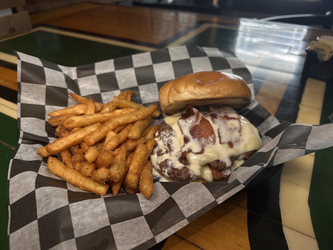 A bacon cheeseburger with melted white cheese and a pile of seasoned fries in a checked paper basket